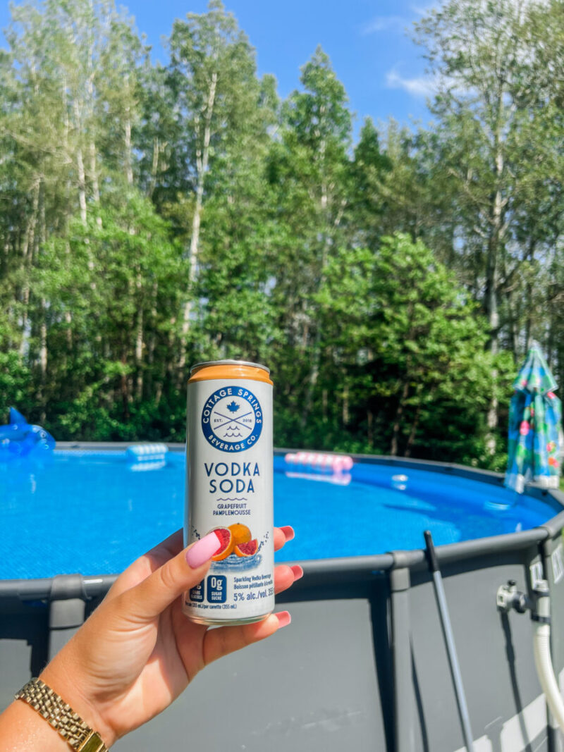 Hand holding a vodka soda cooler in front of our Intex Ultra XTR 24ft above ground pool